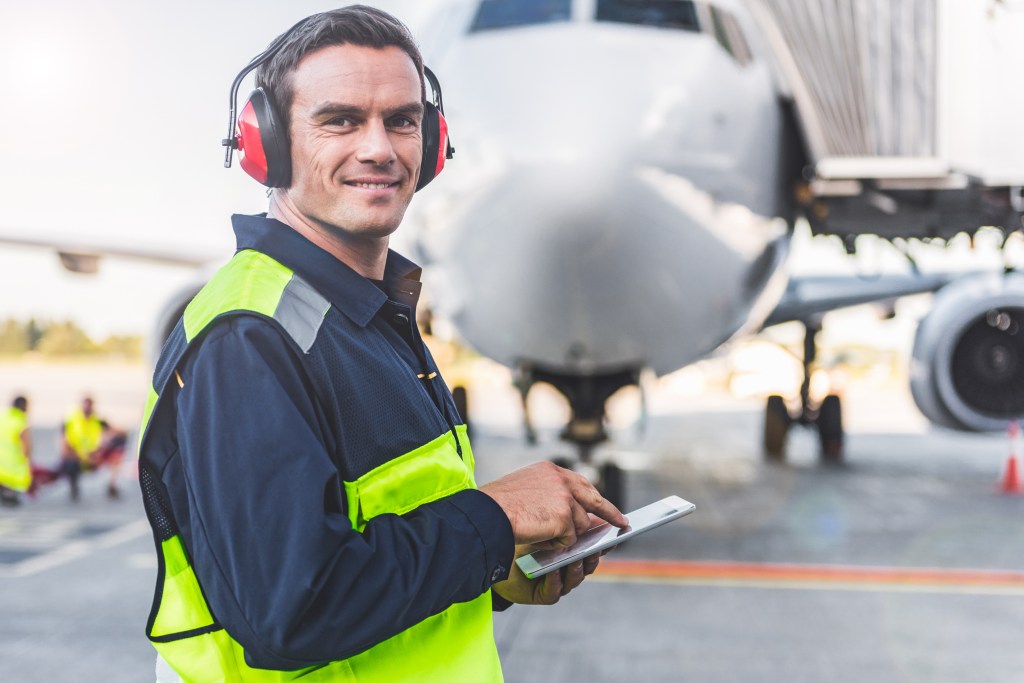 Un employé d'aéroport souriant, portant un gilet de sécurité et des protections auditives, se tient sur le tarmac et utilise une tablette. En arrière-plan se trouve un grand avion de ligne.