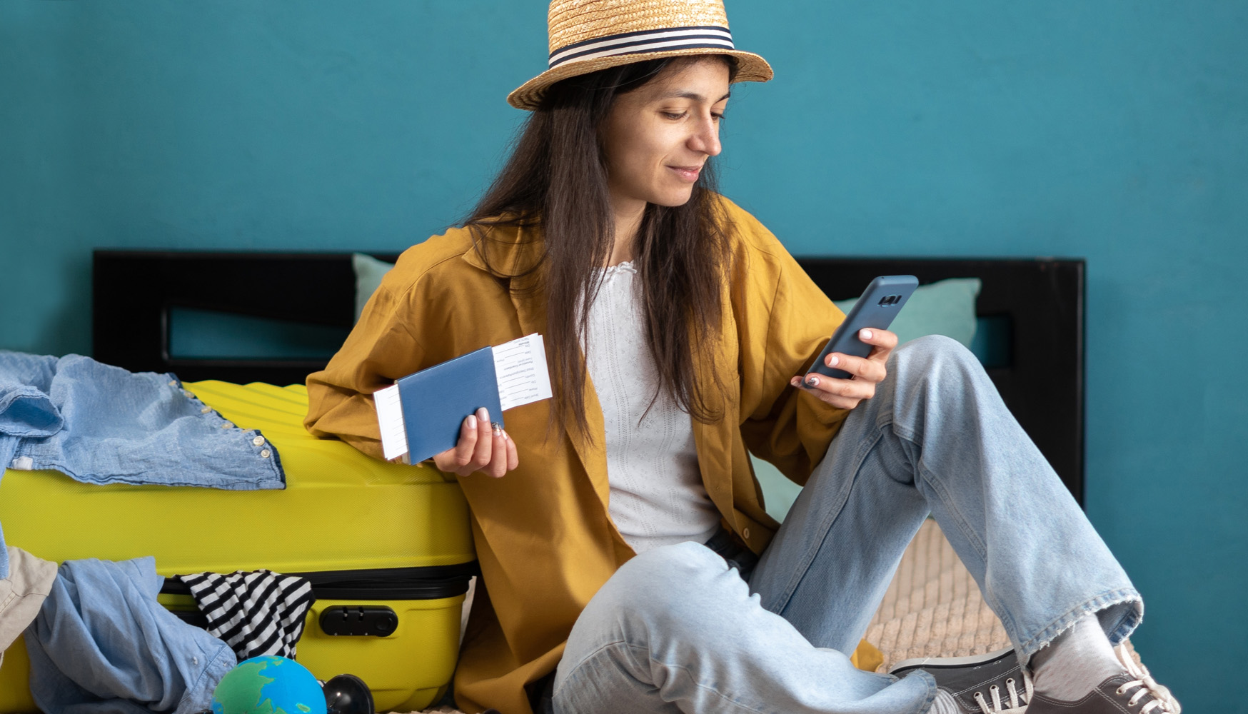 Een lachende vrouw met een strohoed zit op een bed naast een ingepakte gele koffer. Ze houdt een paspoort met vliegtickets in haar hand en kijkt op haar smartphone om haar vakantie te plannen.