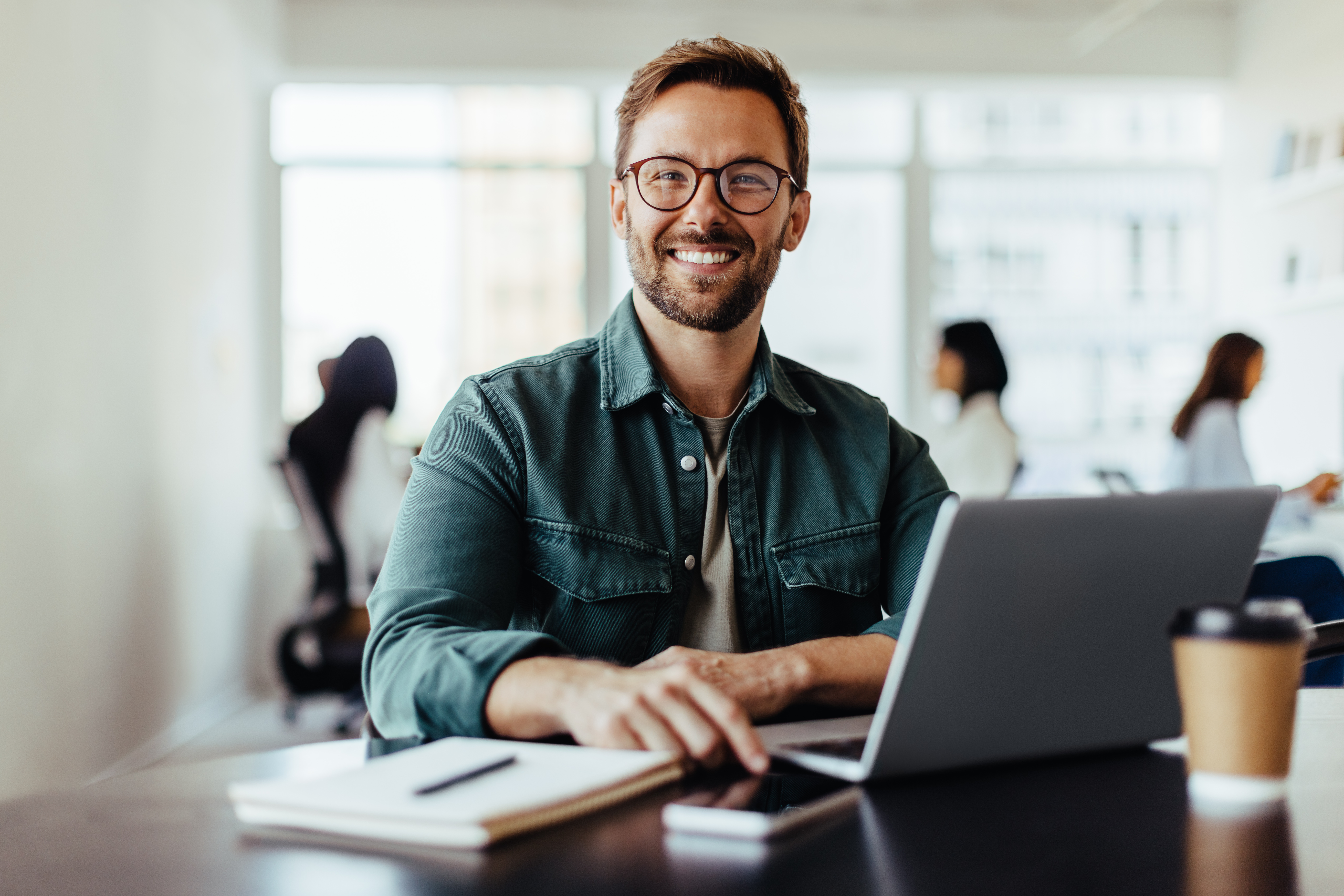 Portret van een lachende zakenman aan het werk in een kantoor, met collega's op de achtergrond in een co-working ruimte.