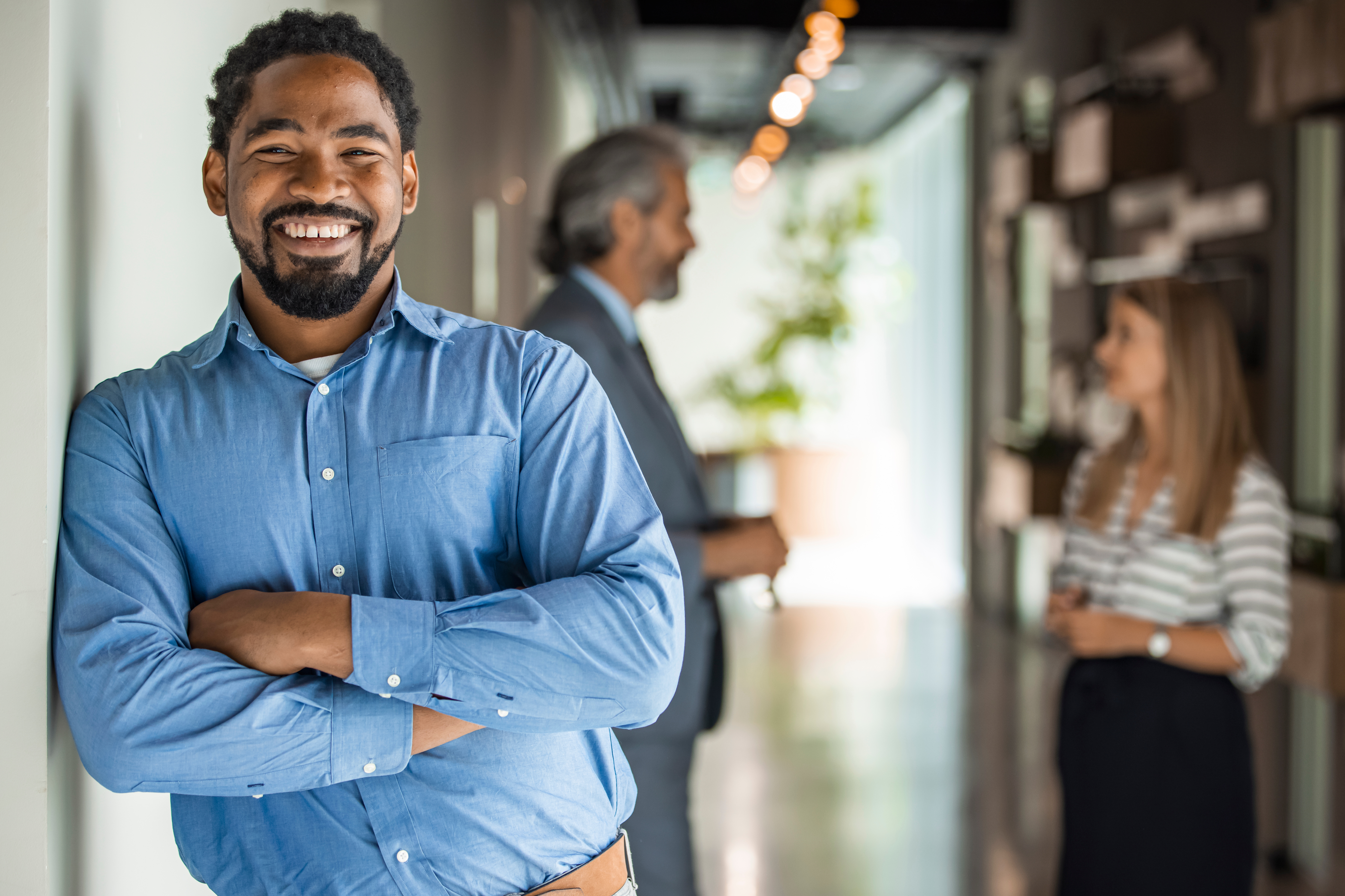 Portrait Of Businessman In Modern Open Plan Office With Business Team Working In Background. African office worker with arms crossed. Successful businessman in modern office